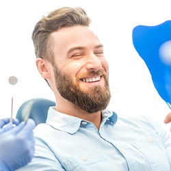 A man with a beard looking at his revised smile in the mirror while at the dentist’s office