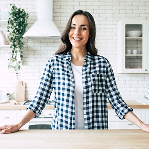 A young woman wearing a plaid button-down and standing in her kitchen smiling after having direct bonding in Homer Glen to repair her minor imperfections